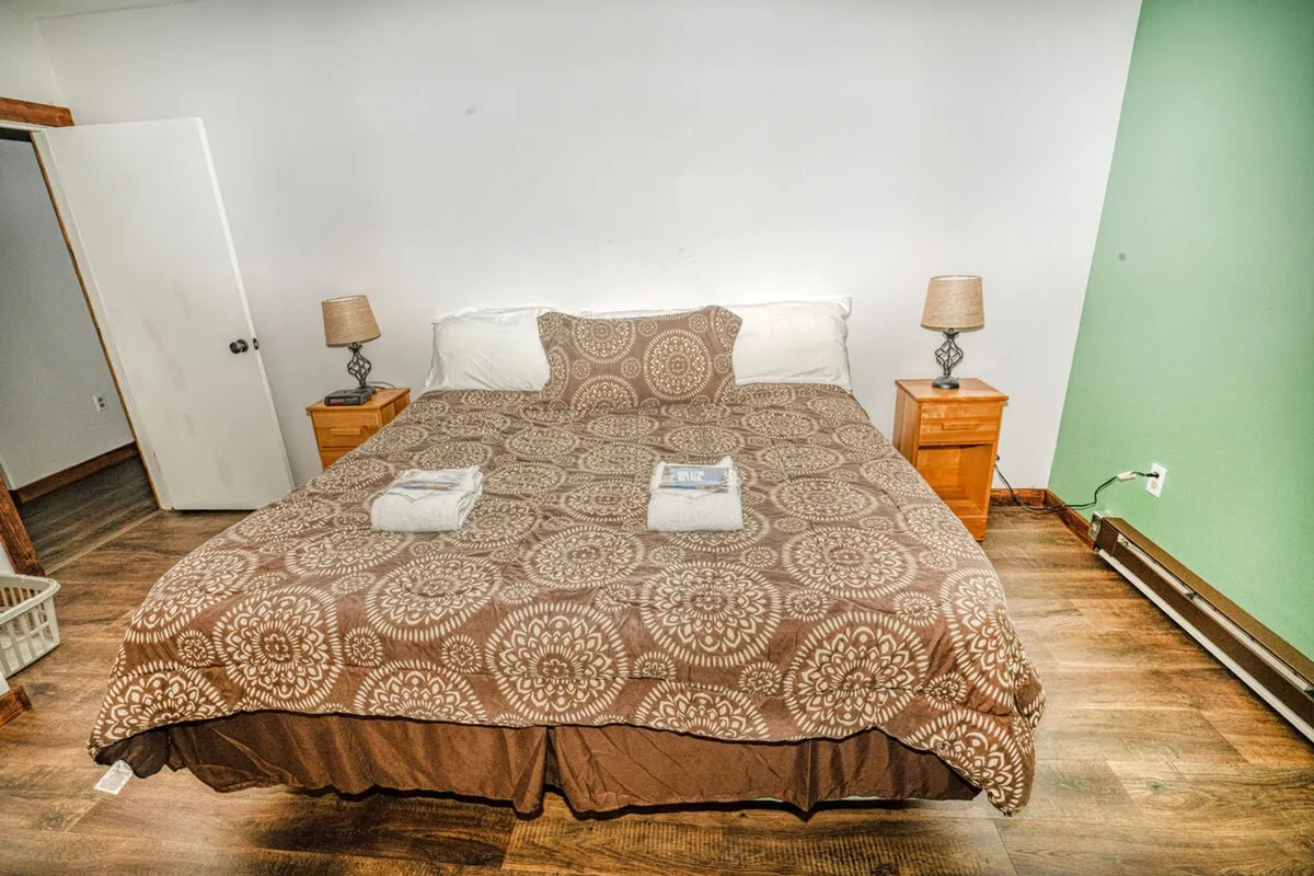 A spacious bedroom featuring a large bed with a patterned cover, flanked by nightstands and lamps, all set against a calming green wall. The ambiance is warm and inviting, suggesting a restful space for families or friends.
