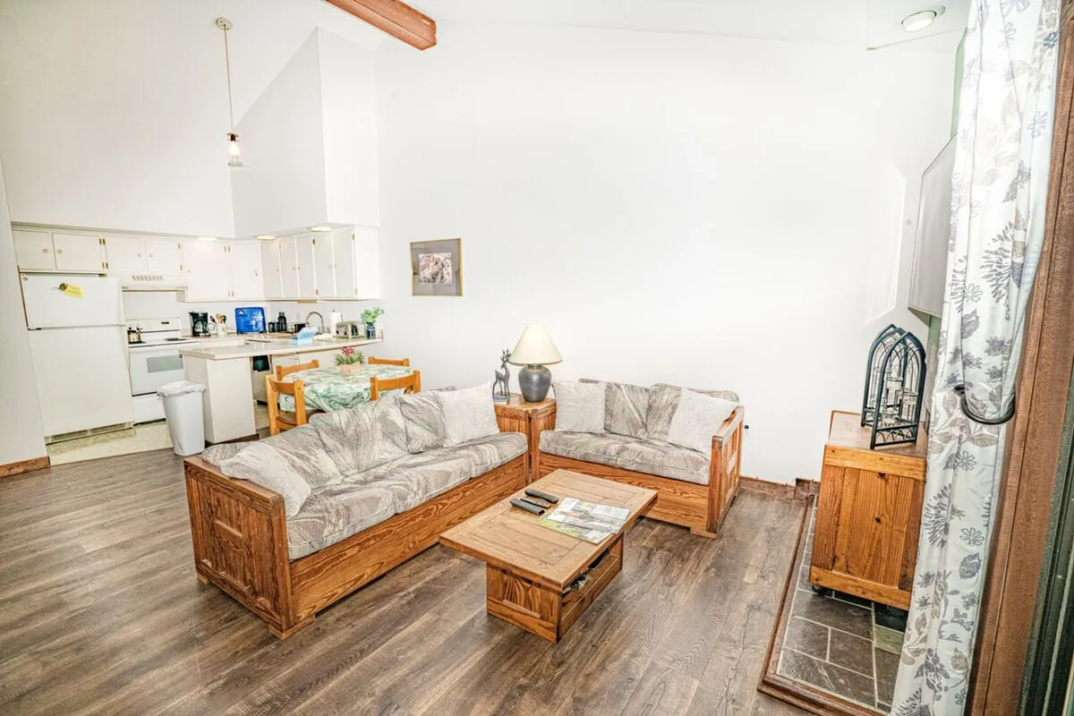 An inviting living space with a sofa set, coffee table, lamp, and partial view into the kitchen area. Morning light fills the room, creating a warm ambiance ideal for relaxing or socializing after a day on the mountains.
